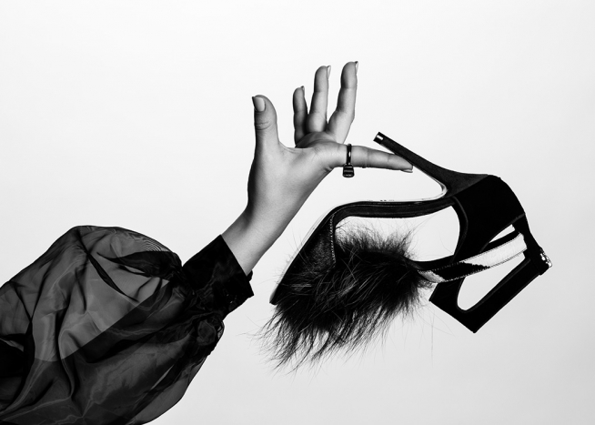  – Schwarz-Weiß-Fotografie eines Schuhs. Der High Heel mit Puschel auf dem Vorderschuh wird von einer Frau in durchscheinender Bluse mit gepufftem Ärmel gehalten.