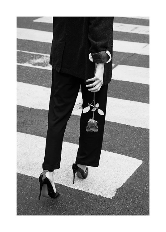  – Schwarz-weiß-Fotografie einer Frau, die einen Anzug und High Heels trägt und mit einer Rose in der Hand einen Zebrastreifen überquert